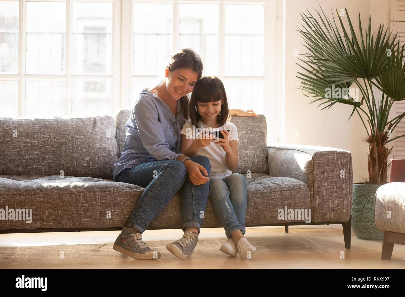 Happy mom and kid daughter using smartphone sitting on couch Stock Photo