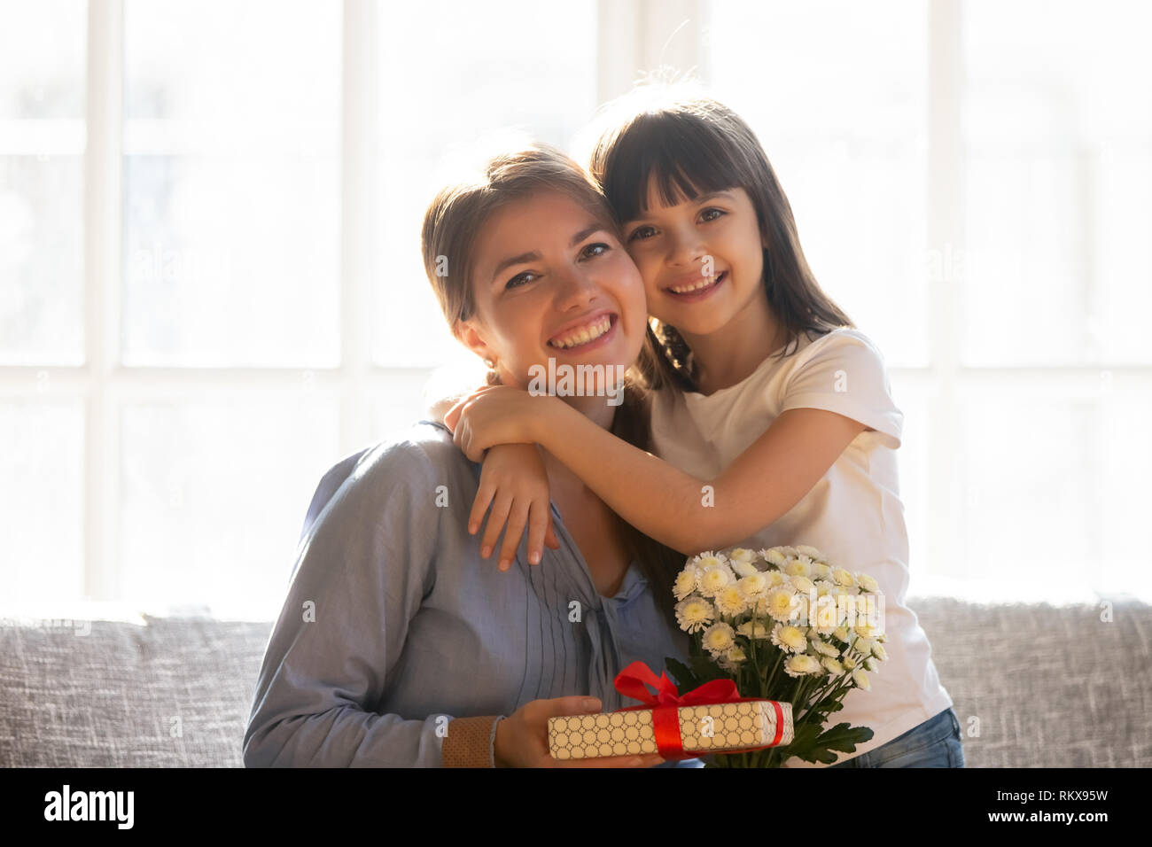 Happy kid daughter embracing mom holding flowers bouquet and gift Stock Photo