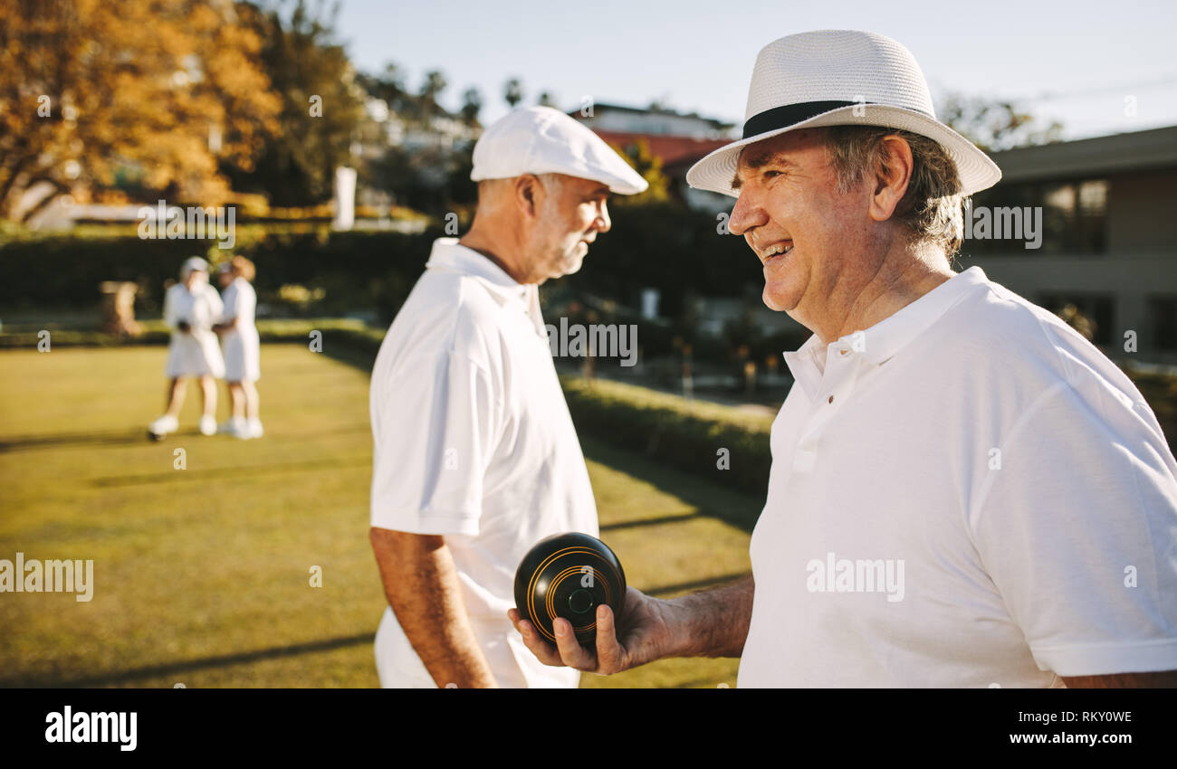 Side view of an old man standing in a park playing boules on a sunny day. Senior man in hat playing boules in a lawn with friends. Stock Photo