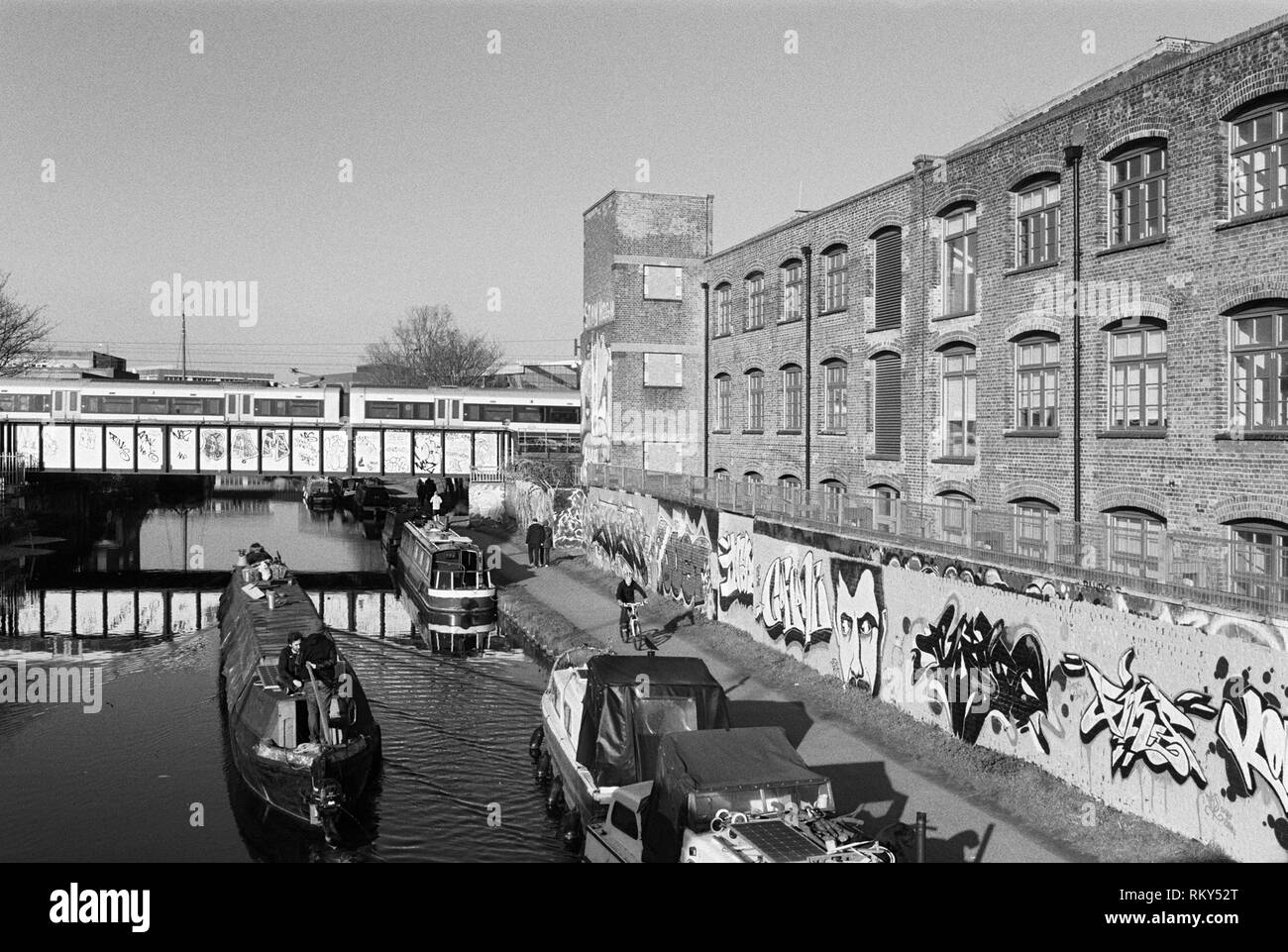 Narrowboat and pedestrians along the River Lea Navigation at Hackney Wick, East London UK, with train crossing over the raiiway bridge in background Stock Photo