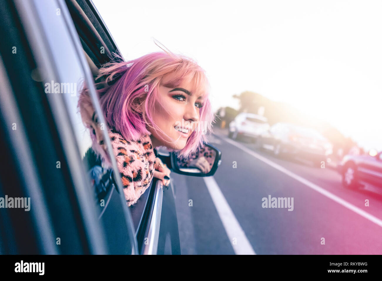 Teen girl with pink hair hangs her head out of a car window on roadtrip. Stock Photo