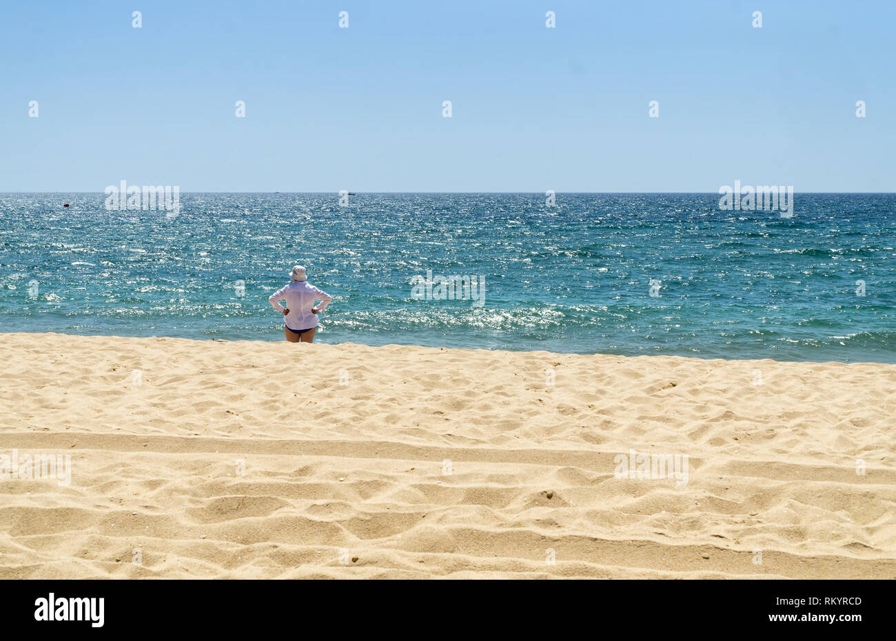 Mature woman in white shirt and white hat is standing on the beach and watching the sea in sunny summer day. Stock Photo