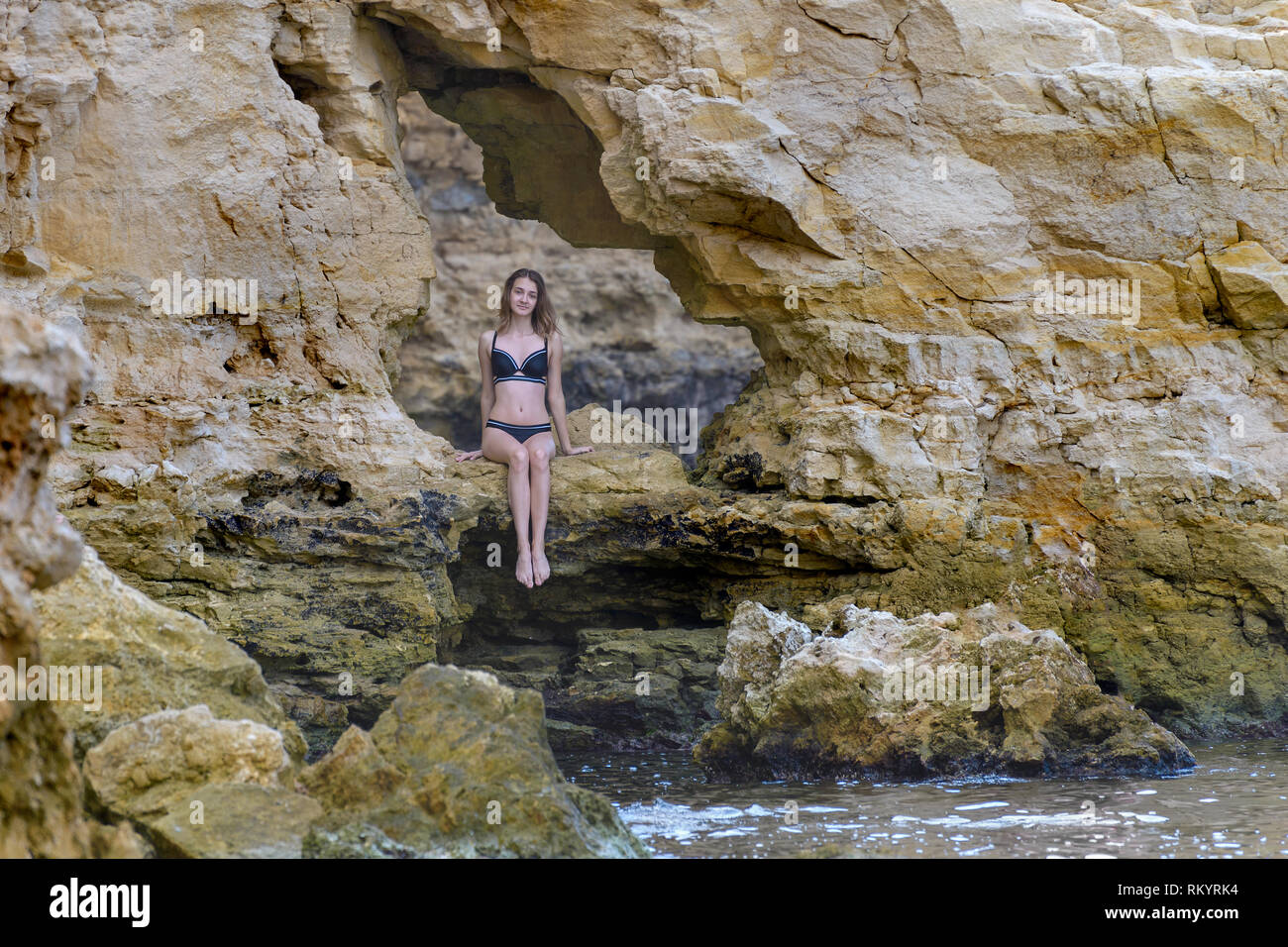 Young girl in black bikini sits in brown rocks by the sea Stock Photo