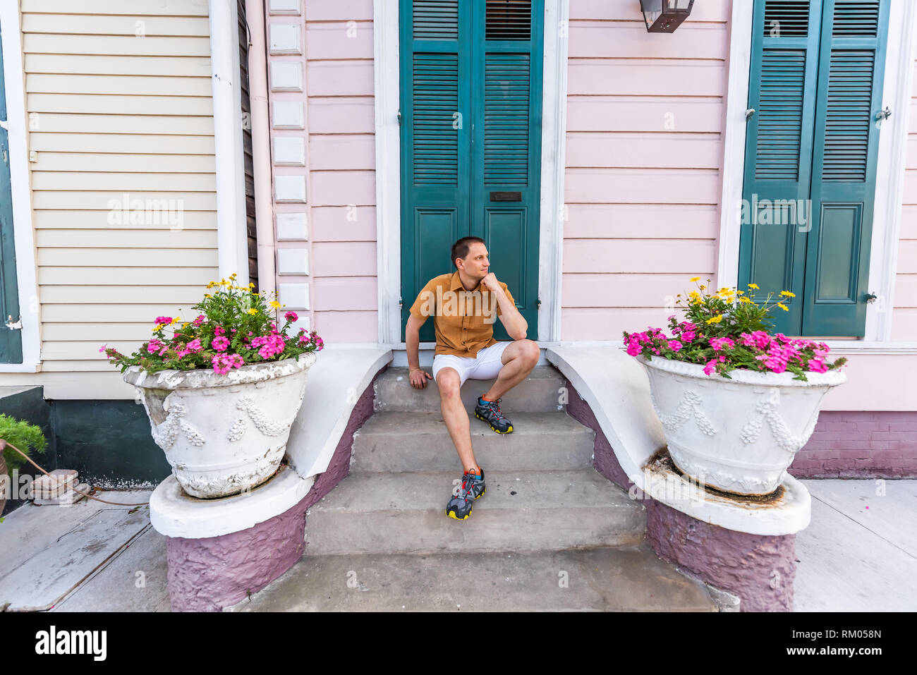 Young local man sitting smiling happy on stairs steps front porch in New Orleans by colorful pink blue green door architecture with yellow flowers in  Stock Photo