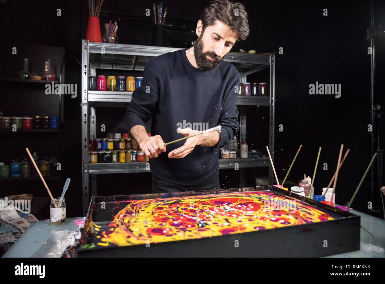 Garip Ay, an Ebru artist, seen in his studio while spreading the Ebru paints with a brush on a tray filled with a mix of water and Tragacanth. Ebru is the art of creating colorful designs by sprinkling and brushing pigments on an aqueous surface to later transfer the drawing to fabric or paper. It is believed that the origin of this technique dates back to the 12th century in Turkistan. The Ottoman calligraphers used Ebru to decorate books, imperial decrees and other documents, thus, Istanbul became the capital of this technique. Stock Photo