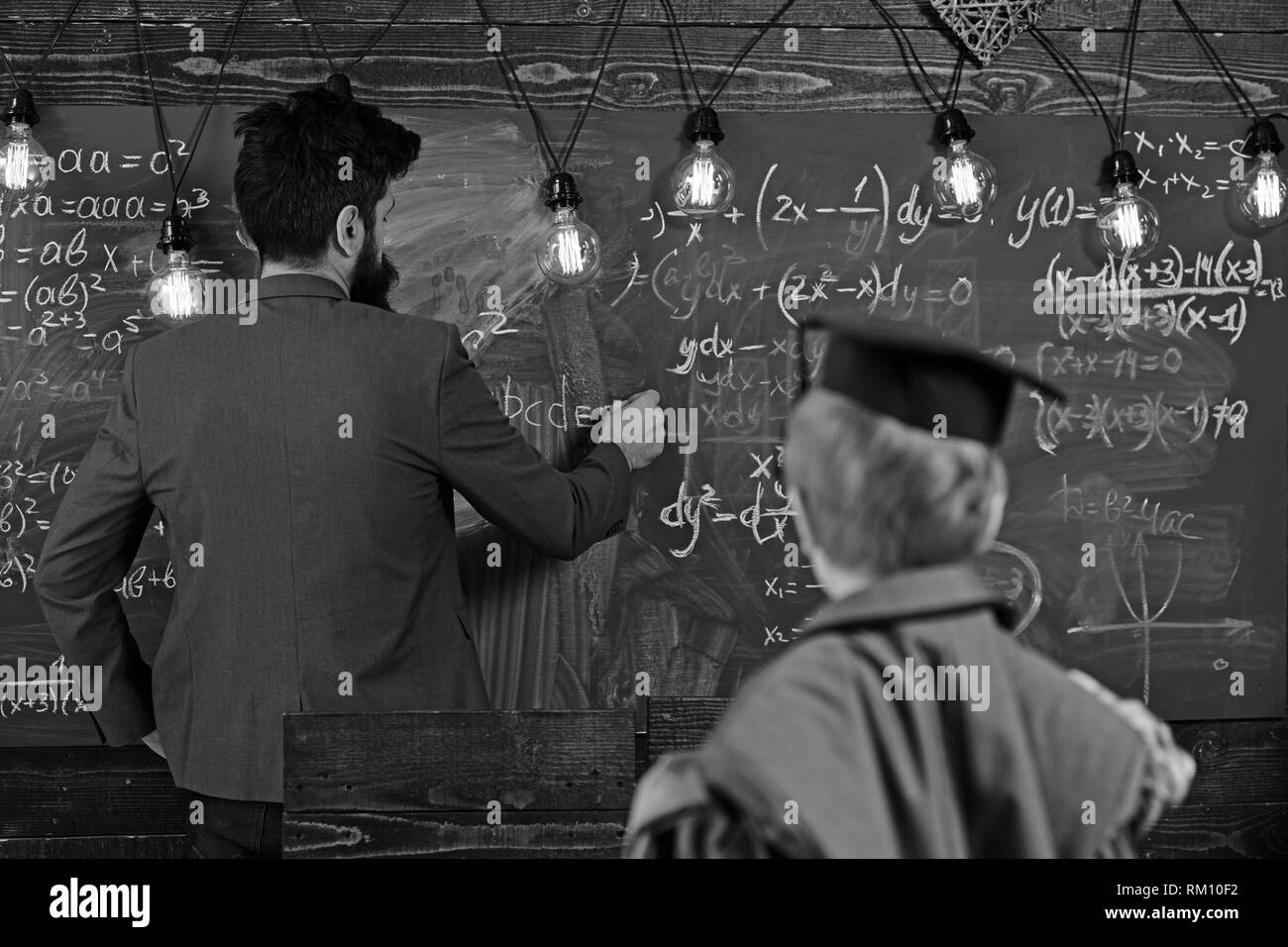 Blurred boy teach man to write on chalkboard. Son and father have school lesson. Parent and child in graduation cap in classroom. Back to school Stock Photo