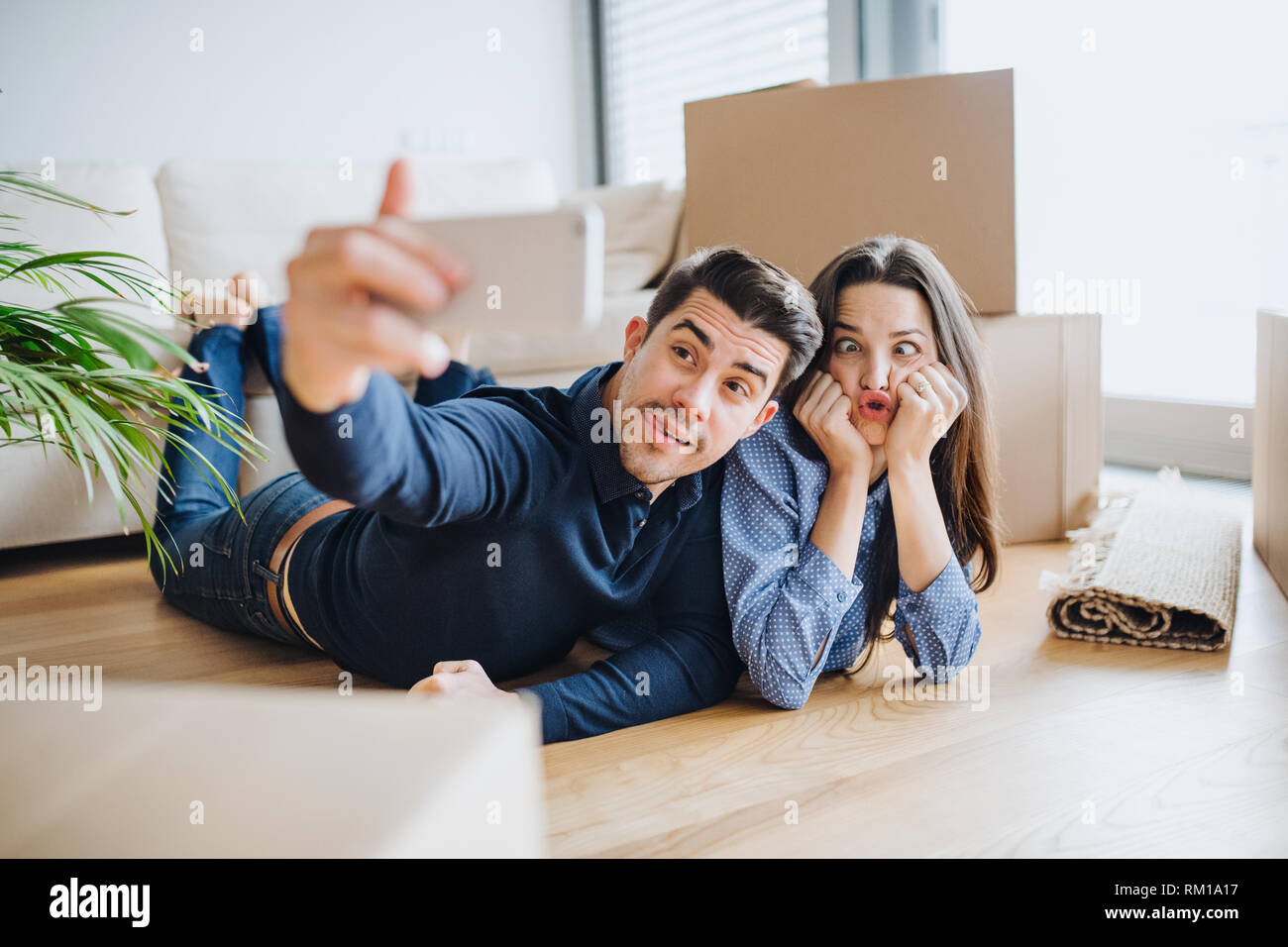A young couple with a smartphone moving in a new home, taking selfie and making a face. Stock Photo