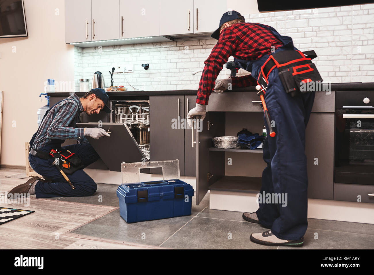 Learning from the best. Two men technician senior and young are sitting near dishwasher with screwdriver in kitchen with instruments Senior man teaches young one Stock Photo