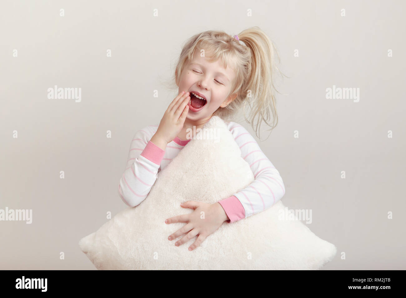 Healthy sleep concept. Cute blonde Caucasian girl child in pink pajamas with closed eyes yawning covering wide open mouth with palm. Sleepy kid with m Stock Photo