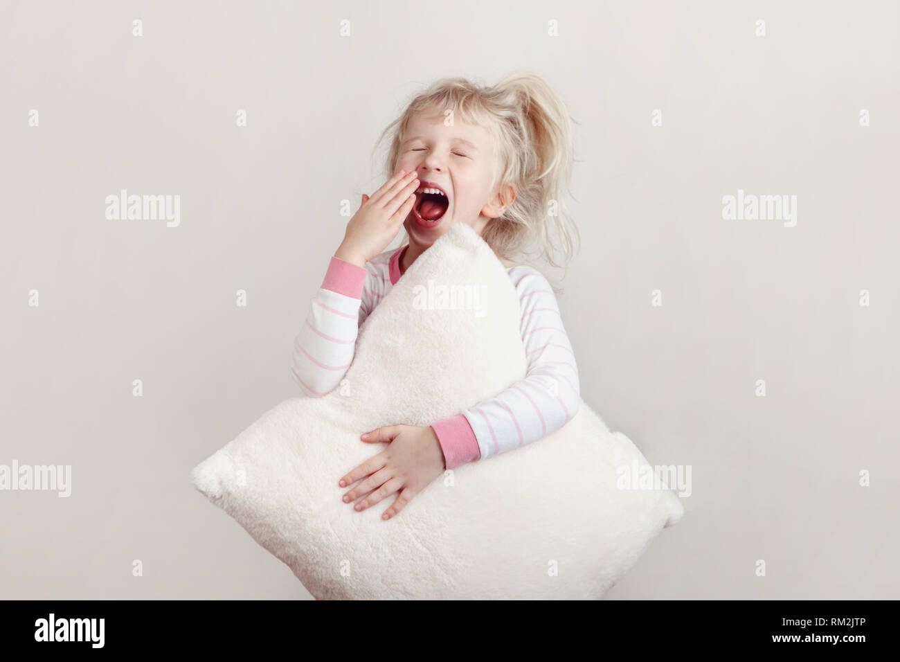 Healthy sleep concept. Cute blonde Caucasian girl child in pink pajamas with closed eyes yawning covering wide open mouth with palm. Sleepy kid with m Stock Photo