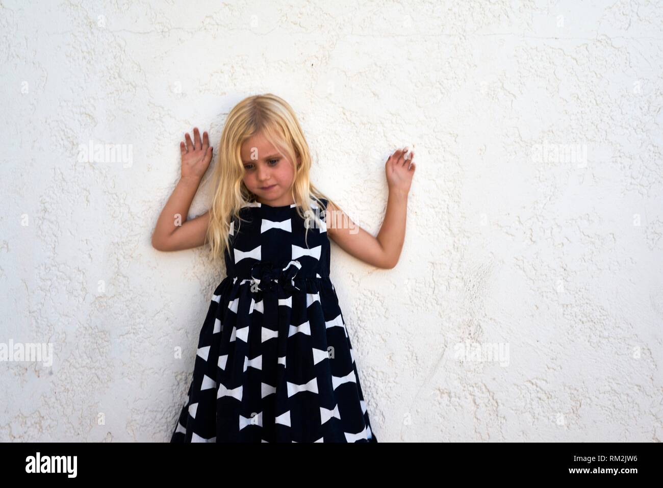 thoughtful female child in dress (metaphor for pressing children in societal norms and gendering) holding arms up, leaning against white wall, Stock Photo