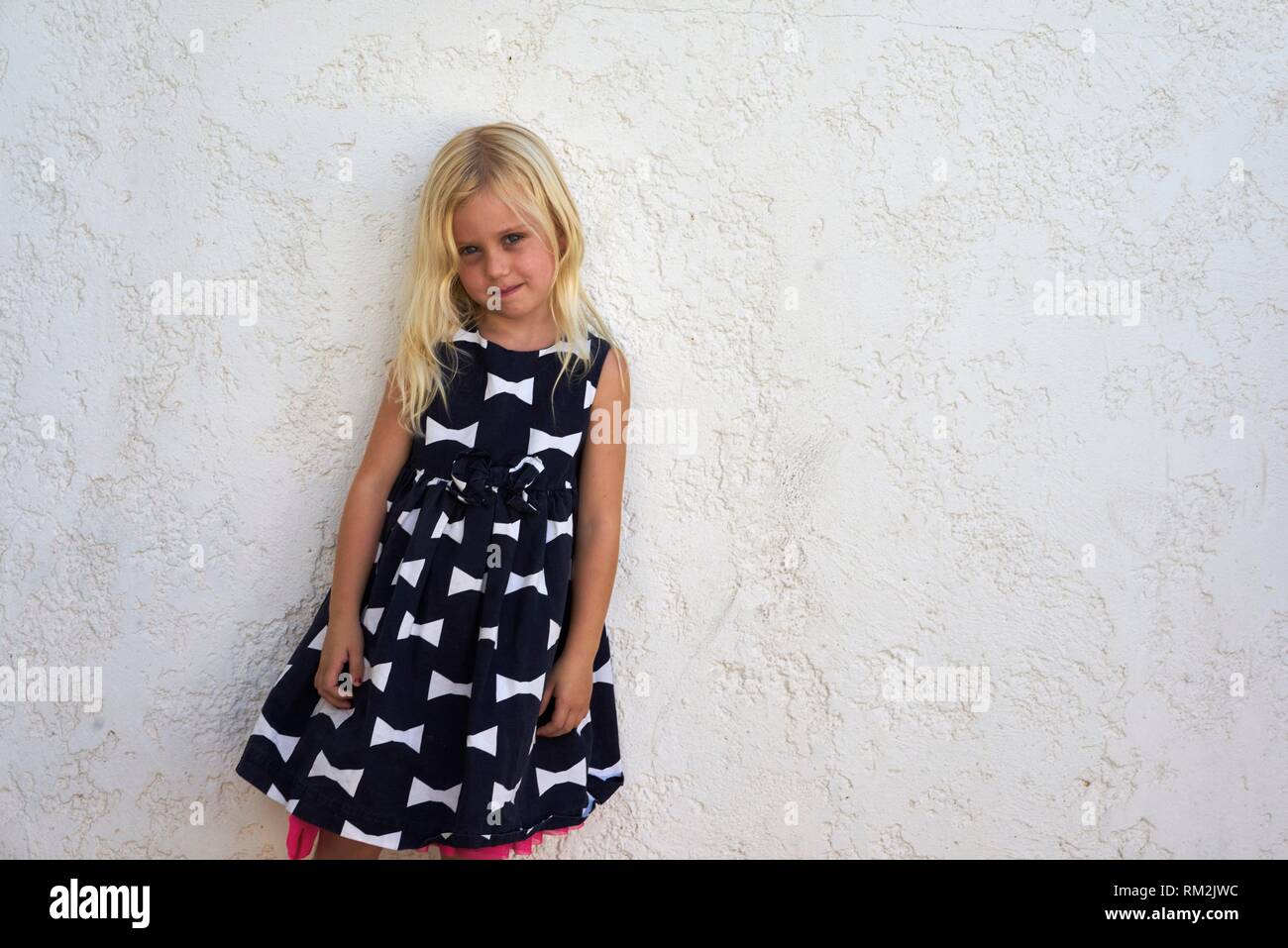thoughtful female child in dress (metaphor for pressing children in societal norms and gendering) leaning against white wall. Australian ethnicity Stock Photo