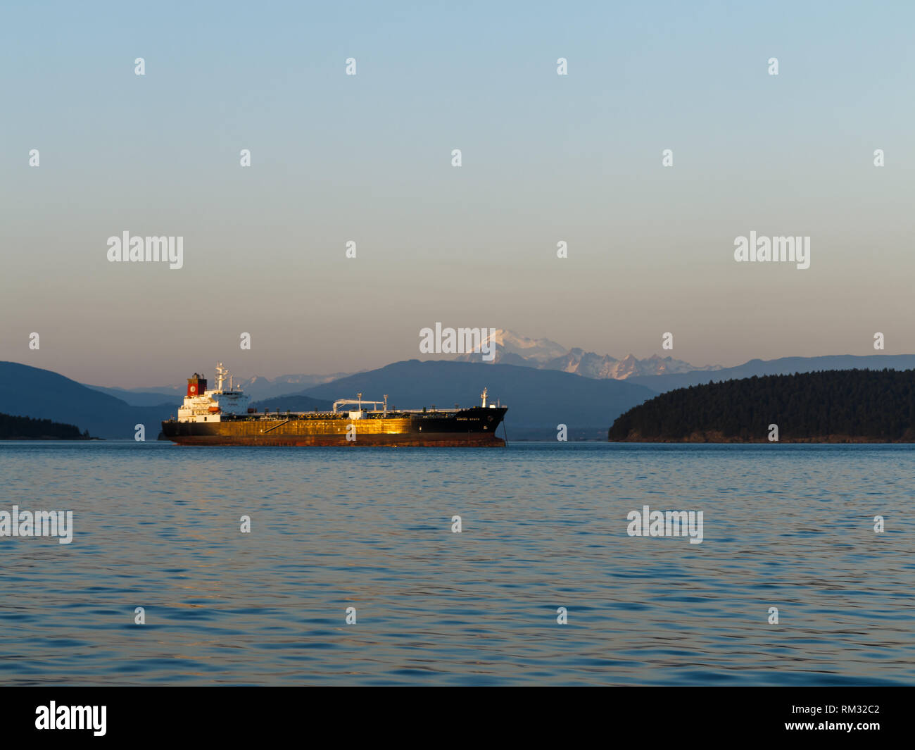 Sun highlights a cargo ship heading out to sea between islands with snow capped mountain peak in background Stock Photo