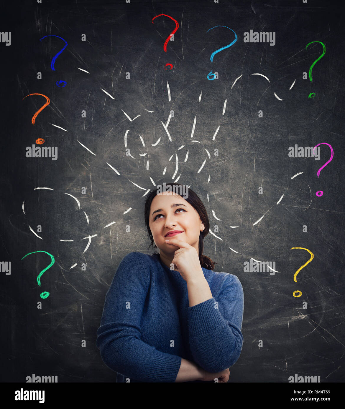 Casual young woman holding hand under chin looking up thoughtfully as a lot of questions as colorful interrogation marks drawn around head over blackb Stock Photo