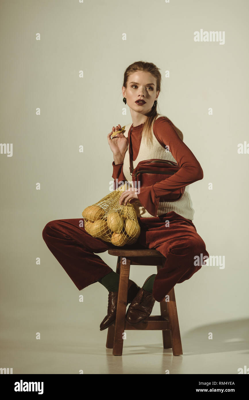 fashionable girl sitting on stool with lemons in string bag on beige Stock Photo