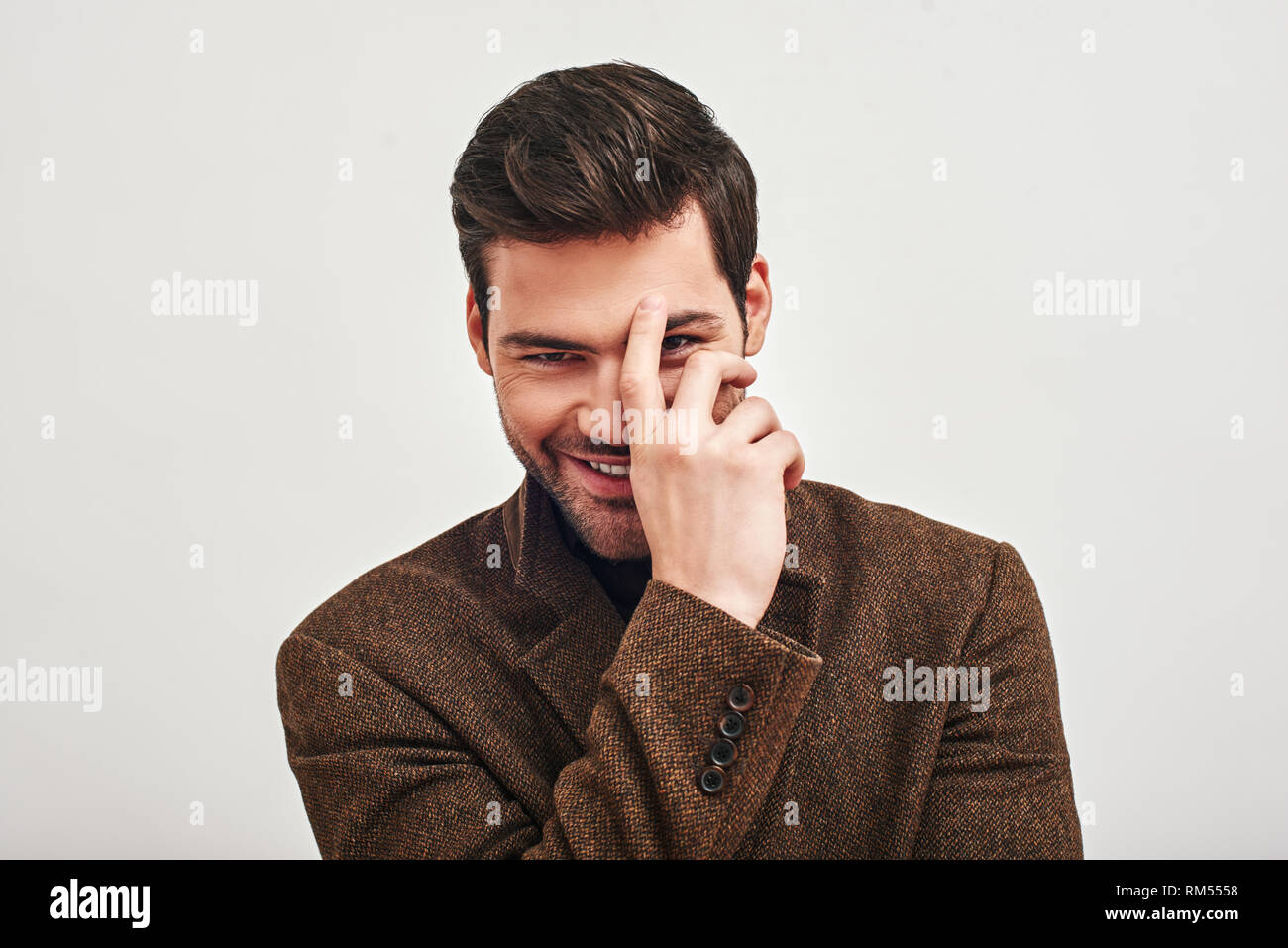 Portrait of stylish dark-haired man wearing brown jacket, touching his forehead, smiling and looking at camera isolated over white background Stock Photo