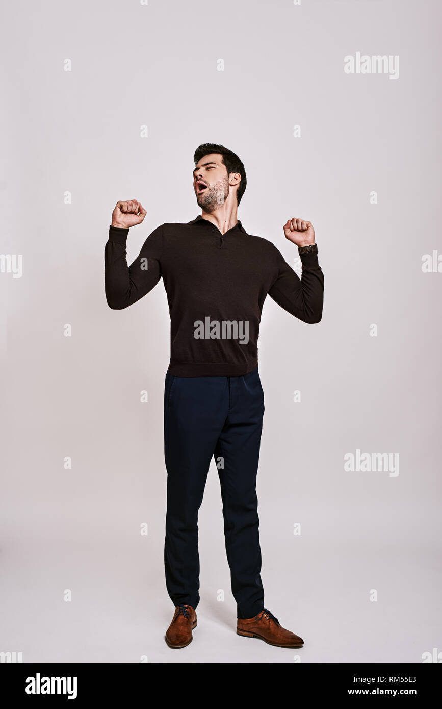 Full-length portrait of stylish dark-haired man wearing blue trousers and brown jumper, standing and yawning isolated over grey background Stock Photo