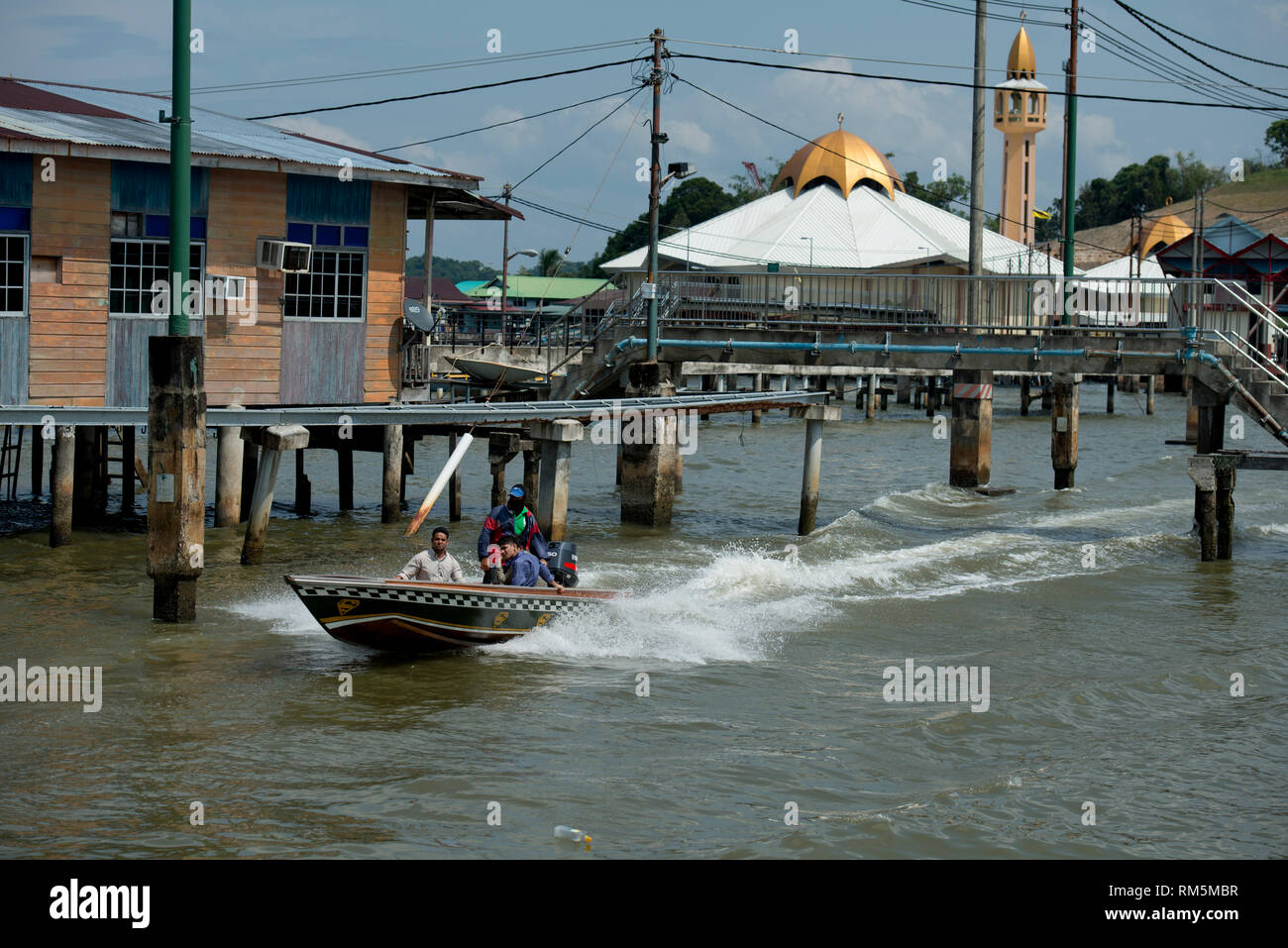 Speedboat water taxi, passing walkway and shacks on stilts in Brunei River with mosque in background, Water Village (Kampong Ayer), Bandar Seri Begawa Stock Photo