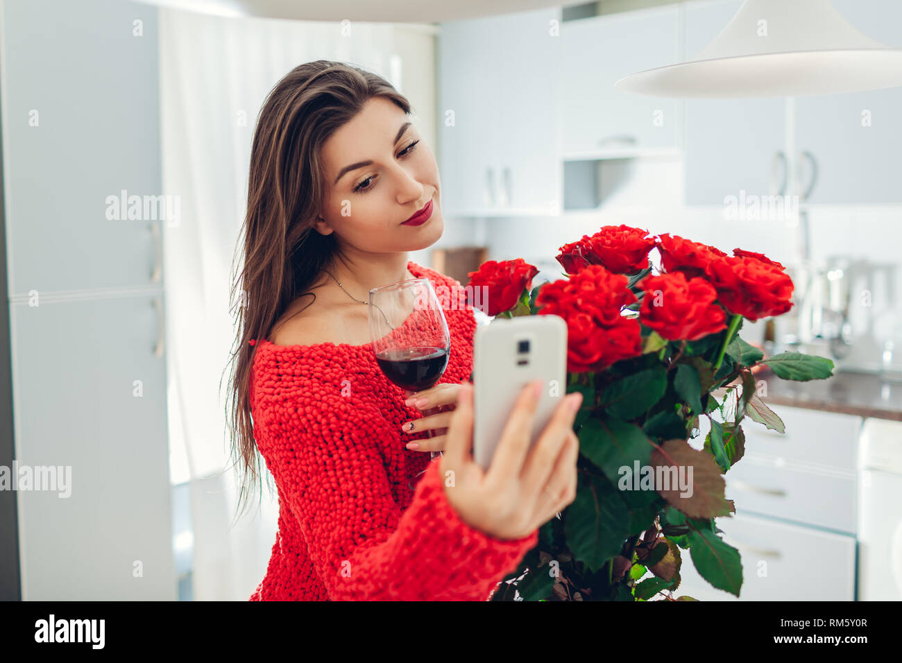 Young woman found bouquet of roses on kitchen. Happy girl taking selfie on phone drinking wine. Valentines day surprise Stock Photo