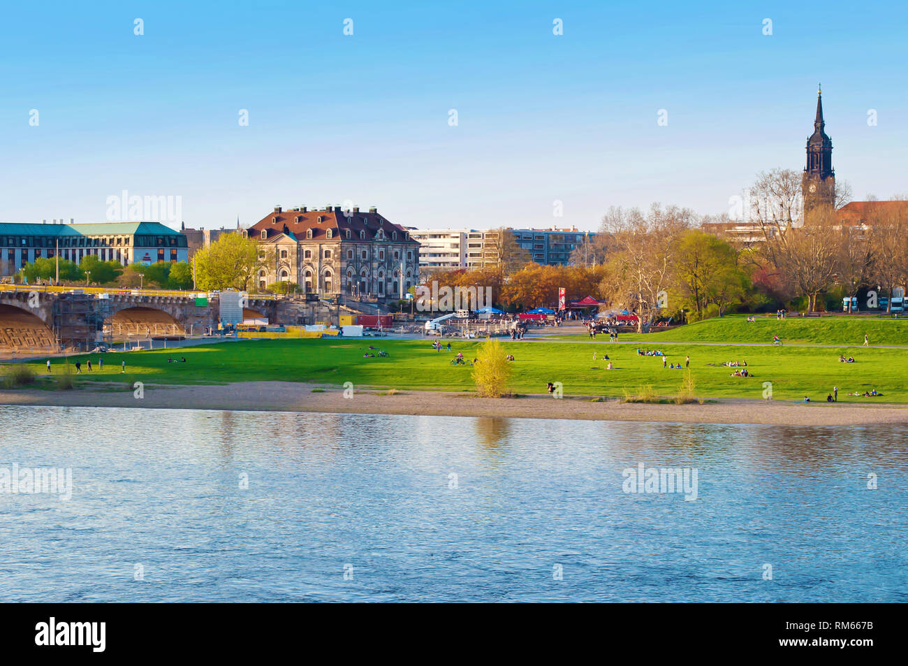 Riverbank of Elbe river in Dresden, Germany. Many people sitting on fresh green lawn and enjoying looking at the shore. Cloudless spring day. Dome of  Stock Photo