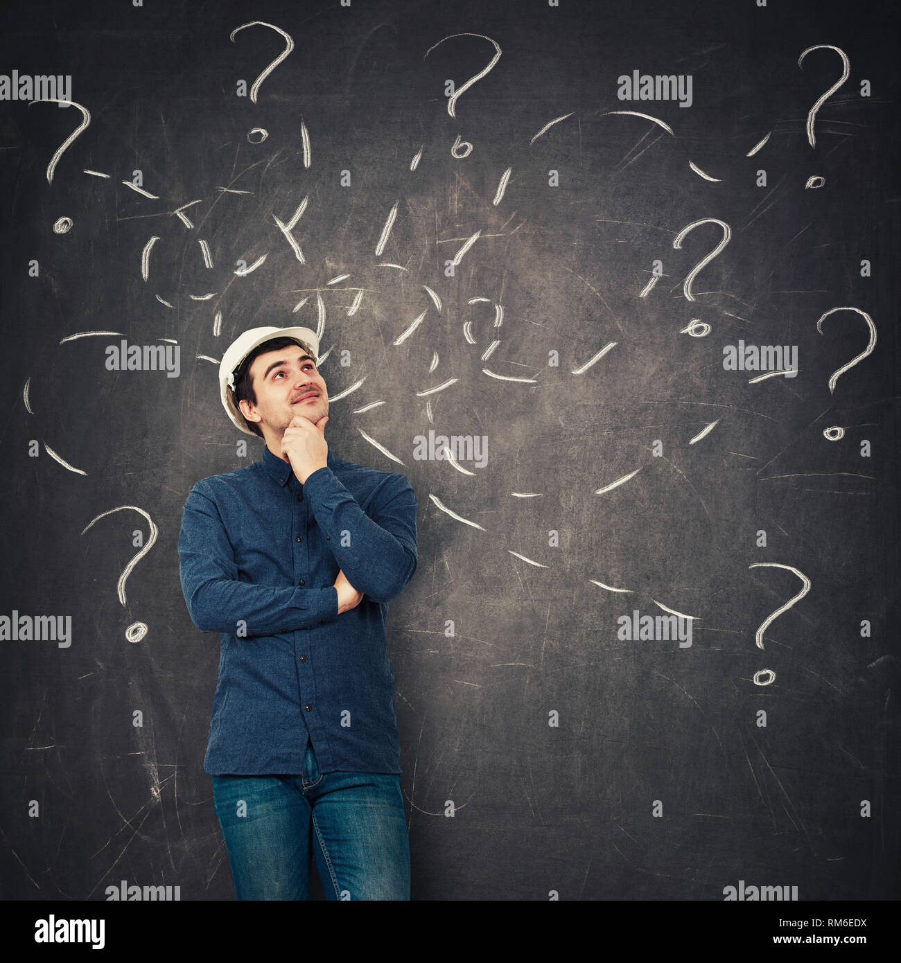 Positive young man engineer wearing protective helmet holding hand under chin looking up thoughtfully in front of a blackboard try to solve different  Stock Photo