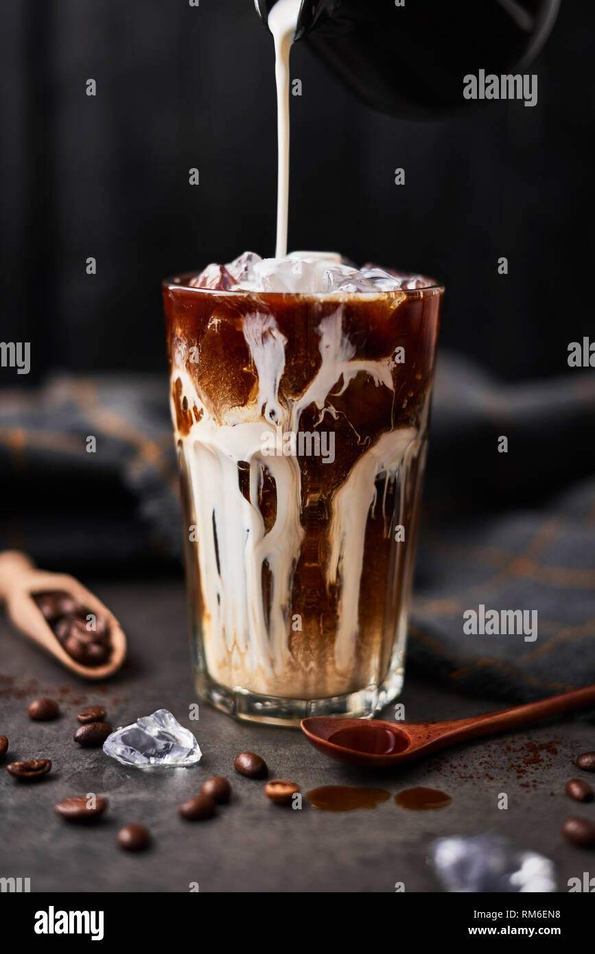 Ice coffee in a tall glass with cream poured over and coffee beans on dark concrete table over black wooden background. Cold summer drink. Copy space  Stock Photo