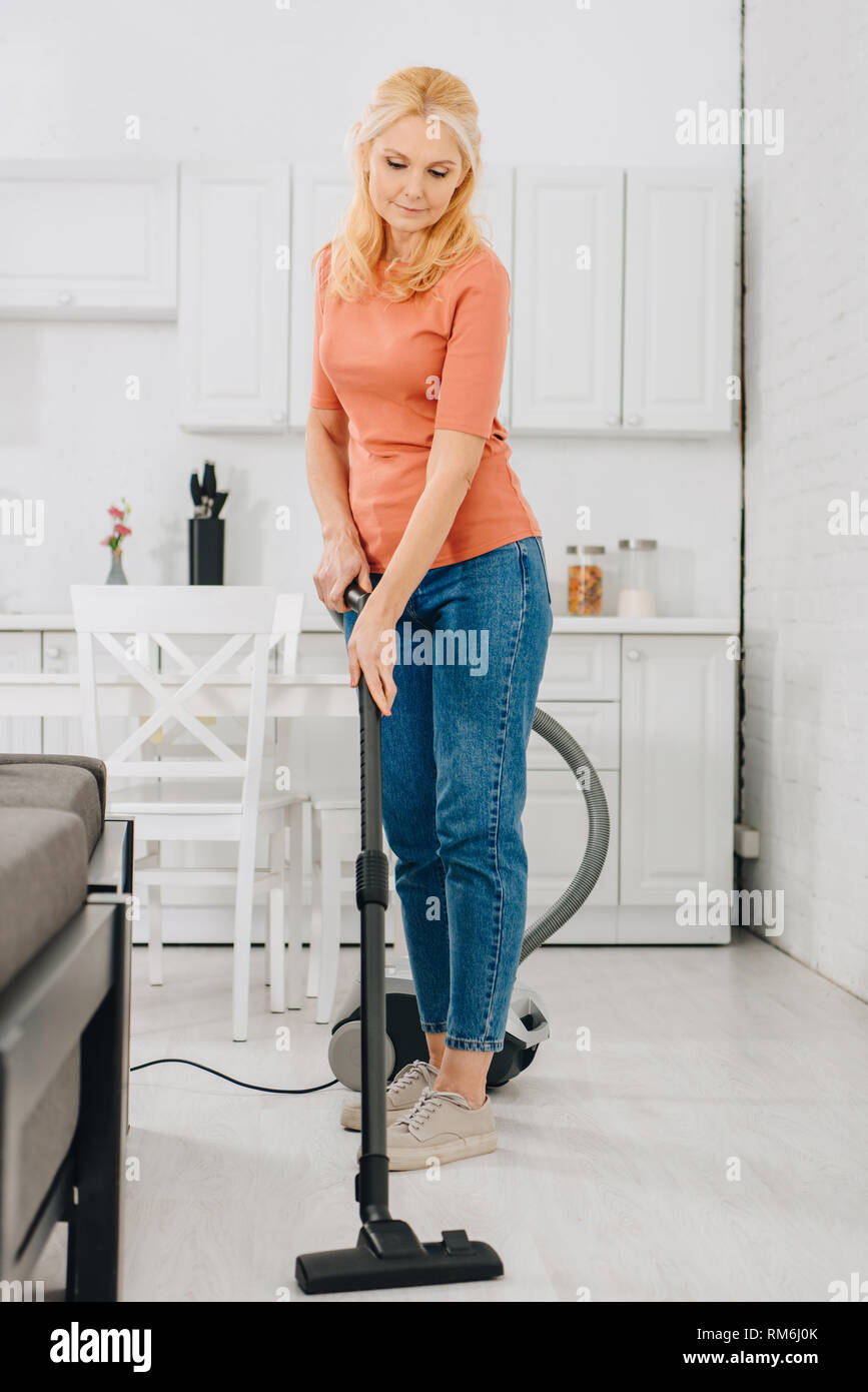 Senior woman in jeans cleaning floor with vacuum cleaner Stock Photo