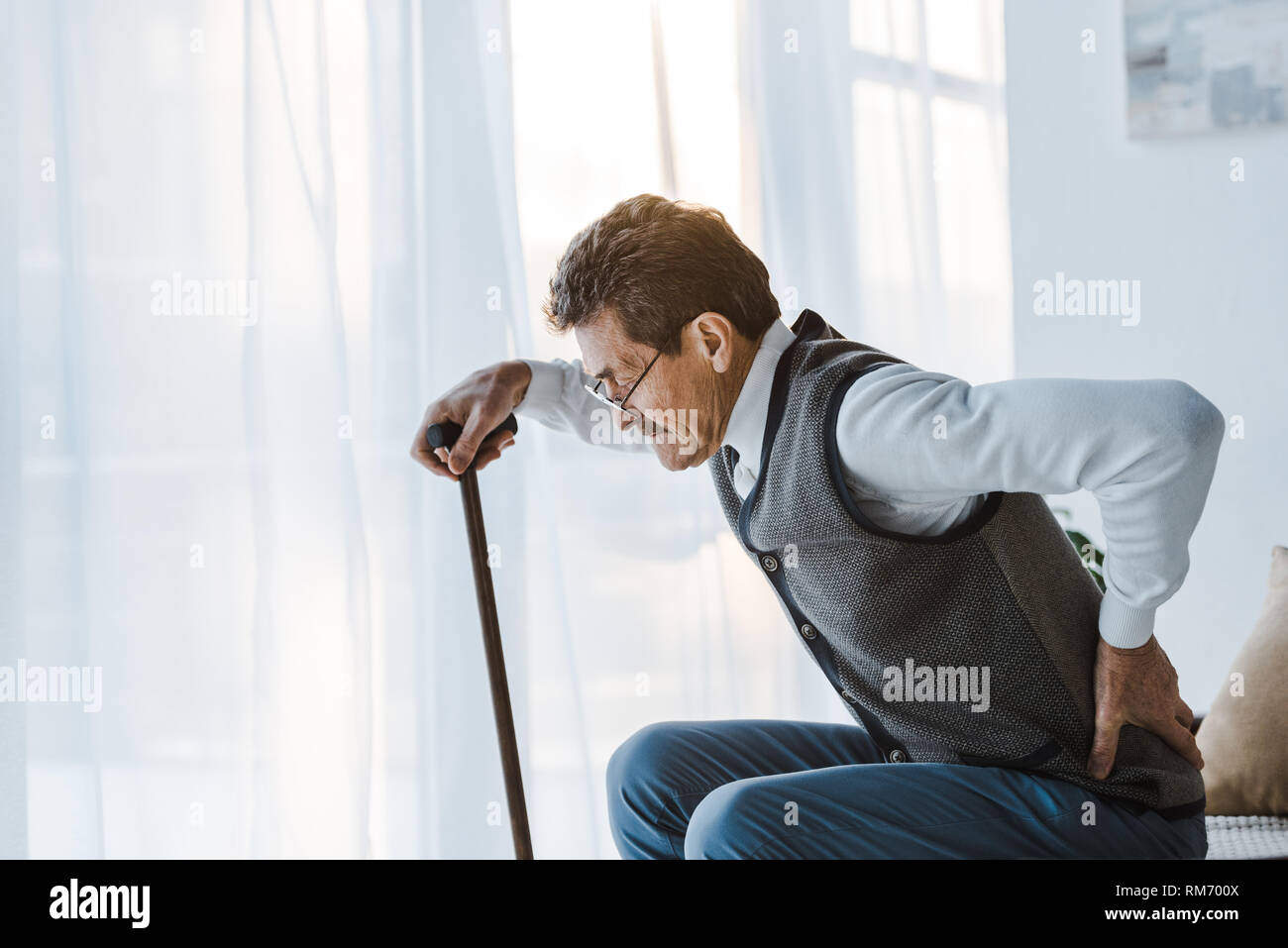 senior man with walking cane holding back while standing up from sofa ...