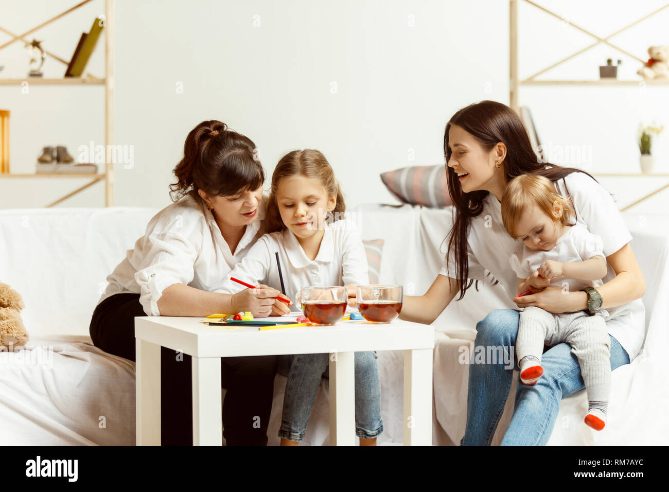 Two little girls their attractive young mother and their charming grandmother sitting on sofa and spending time together at home. Generation of women. International Women's Day. Happy Mother's Day. Stock Photo