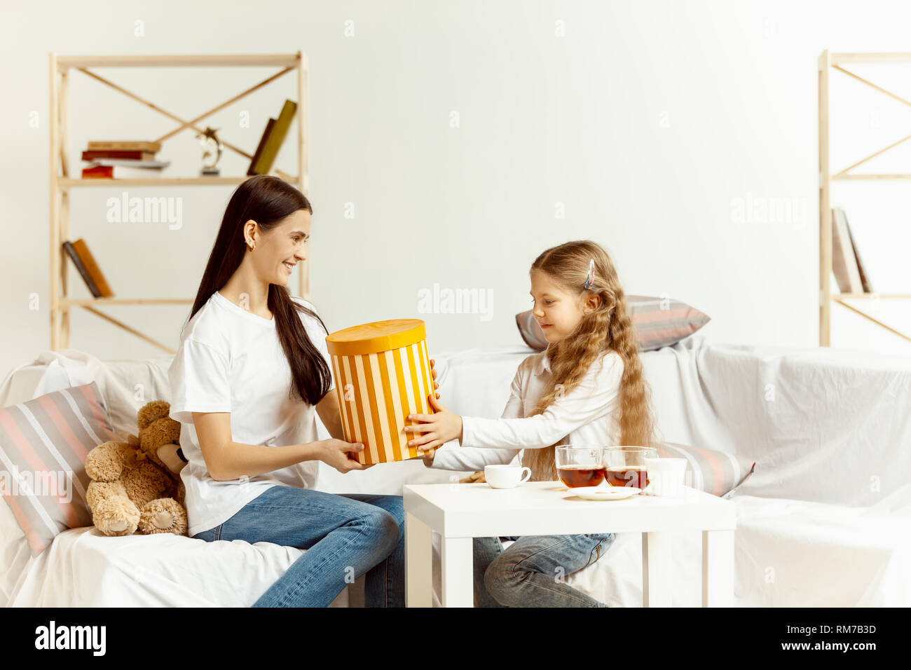 Little girl and her attractive young mother sitting on sofa with gift and spending time together at home. Generation of women. International Women's Day. Happy Mother's Day. Stock Photo