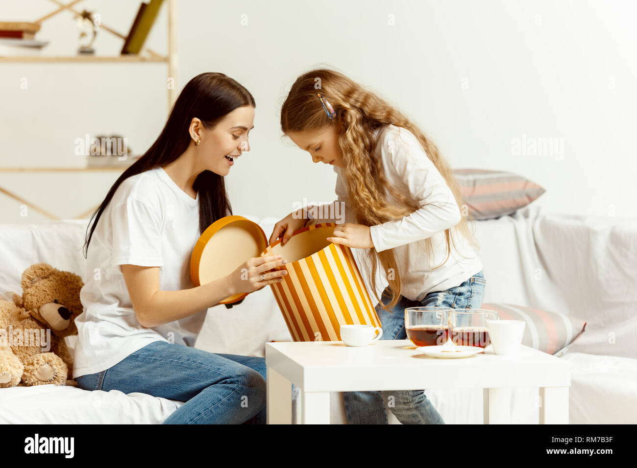Little girl and her attractive young mother sitting on sofa with gift and spending time together at home. Generation of women. International Women's Day. Happy Mother's Day. Stock Photo
