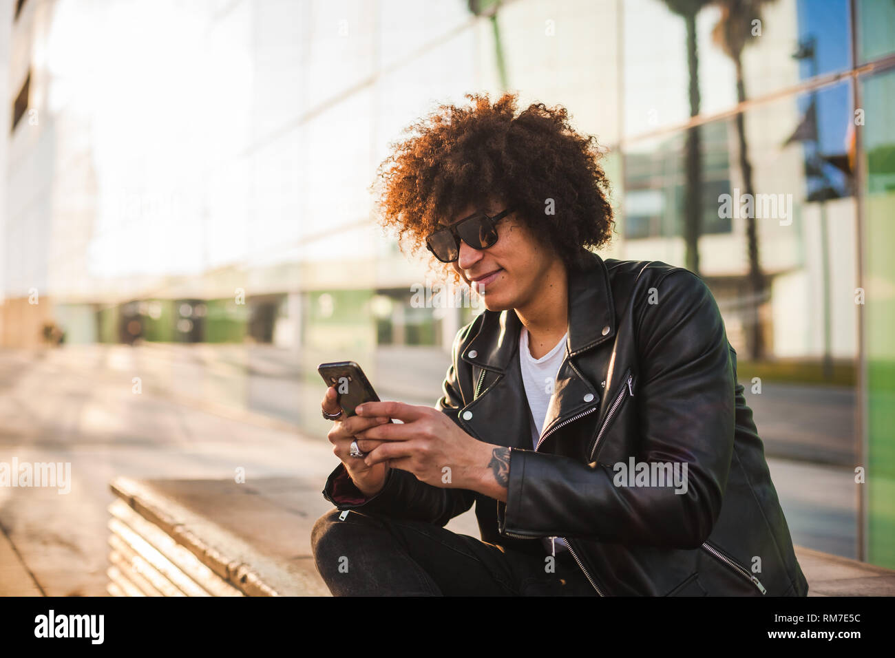 A handsome black man with modern afro hairstyle sitting relaxed in the street using a smart phone Stock Photo