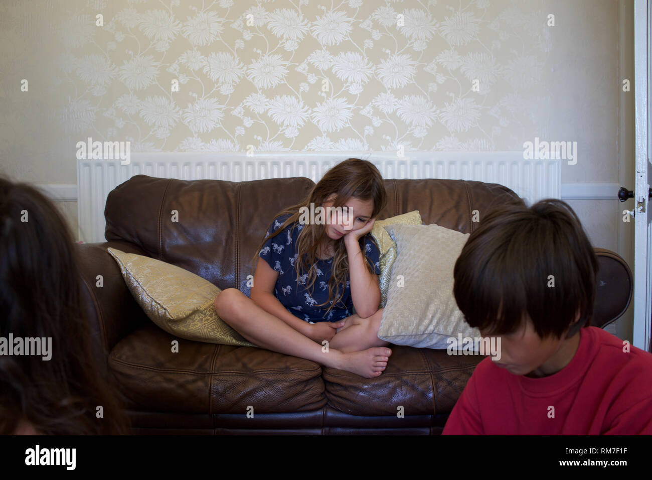 Bored looking child sits cross-legged on sofa, with older child(ren) in foreground. Rugby, England Stock Photo