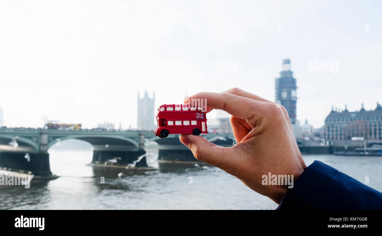the hand of a caucasian man holding a miniature of a red double-decker bus, typical of London, United Kingdom, with the Westminster bridge and the the Stock Photo