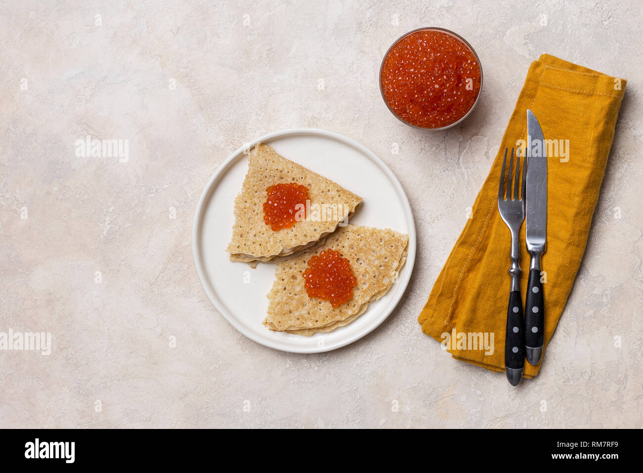 Festive pancakes with red caviar on white plate with fork, knife and napkin, jar of caviar, on white background. Concept of festive food. View from ab Stock Photo