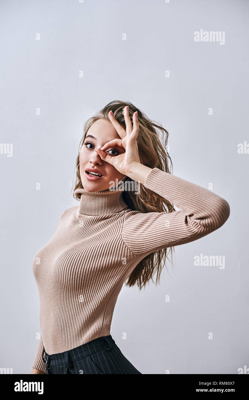 Portrait of young beautiful girl with brown hair, wearing beige turtleneck making OK sign and looking at camera isolated over white background Stock Photo