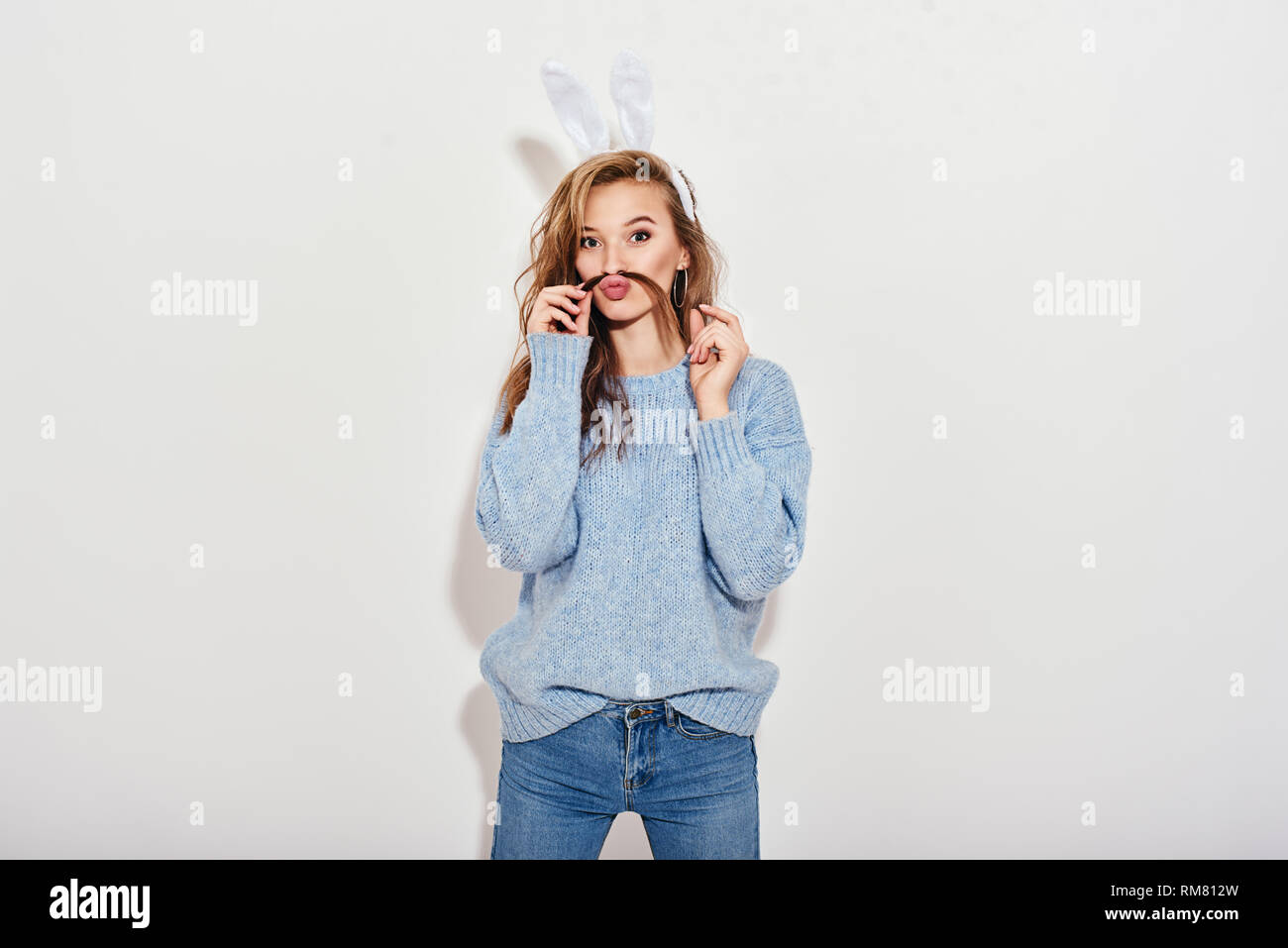 Portrait of cute playful girl wearing blue sweater, jeans and bunny ears  holding a strand of her hair above the lip, looking at camera with widened  eyes, amazedly, isolated over white background