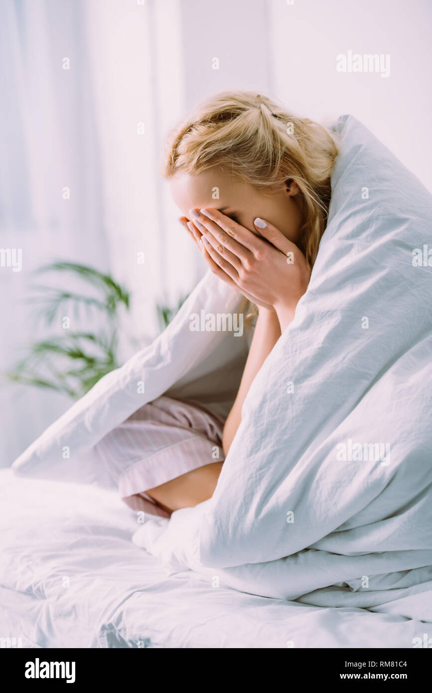 woman in blanket covering face with hands and crying in bed at home