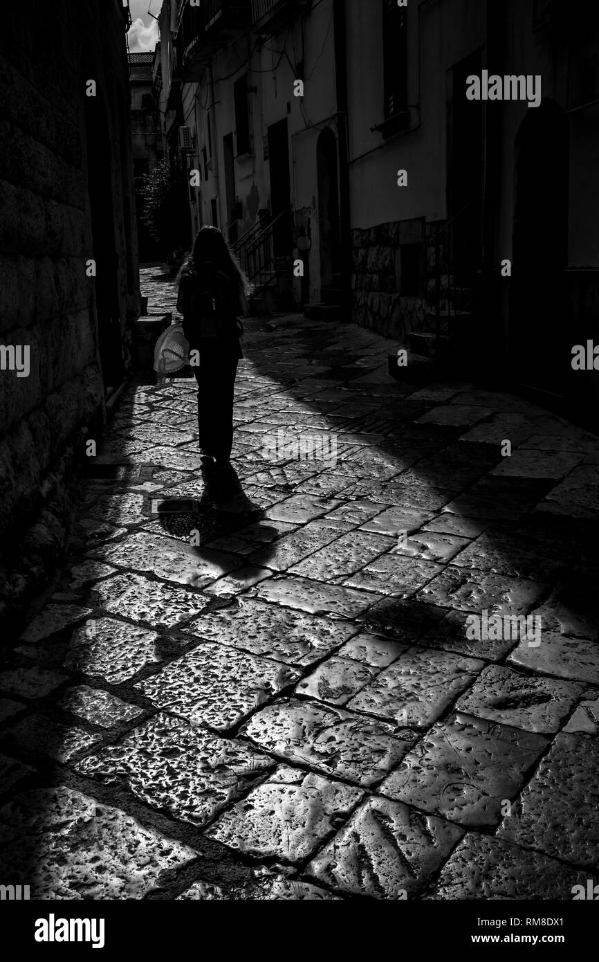 Sun-lit stone narrow street with beautiful girl walking silhouette in Altamura, Apulia, Italy. Black and white high contrast photograph taken against the Sun light in sunny summer afternoon Stock Photo