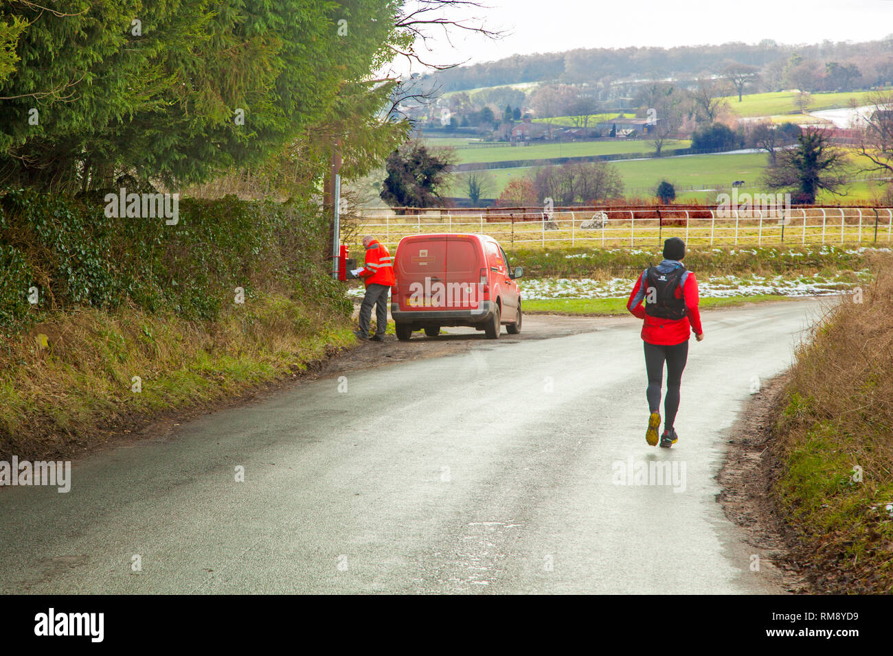 Postman wearing  high vis jacket in post van making a Rural letter collection from a post box in Cheshire countryside with a jogger running past Stock Photo