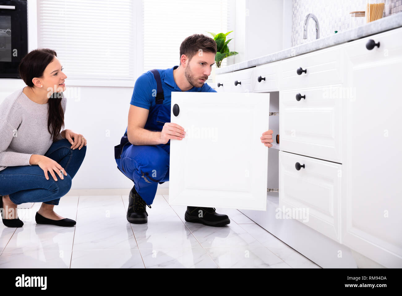 Woman Looking At Male Handyman Installing Cabinet Door In The Kitchen Stock Photo