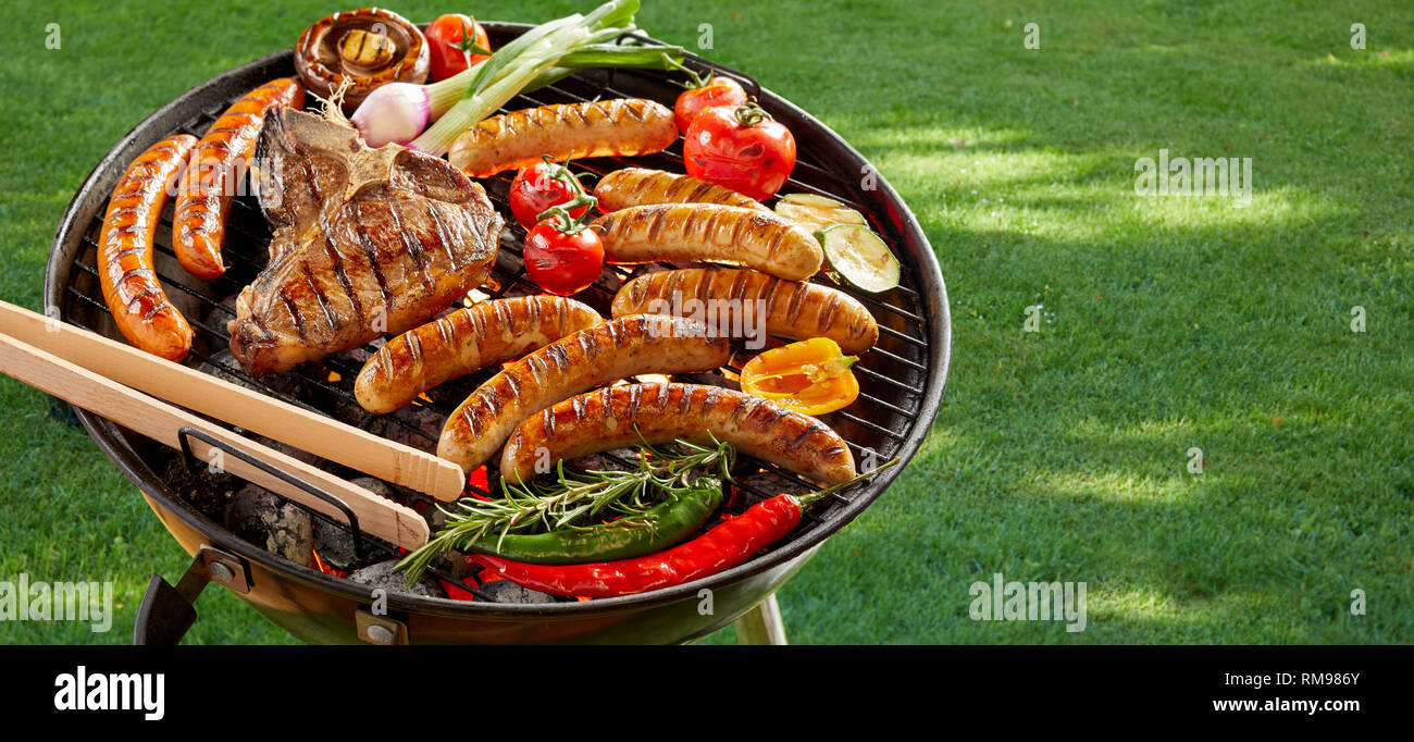Meat and vegetables grilling on an outdoor portable barbecue on fresh green summer grass with pork sausages, beef steak, bell peppers, chili, onion, z Stock Photo