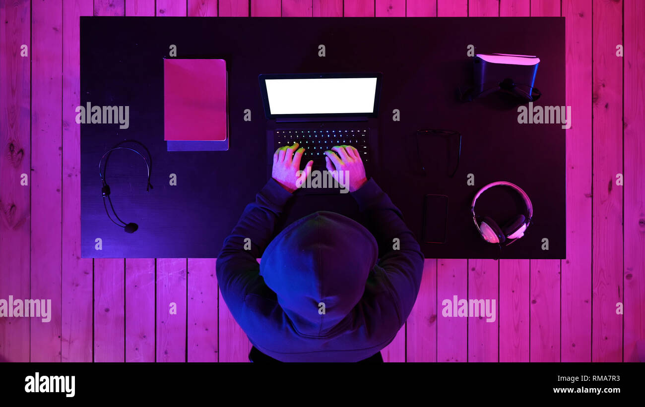 Hacker in hoodie typing code in program using laptop in dark studio. White Display. Stock Photo