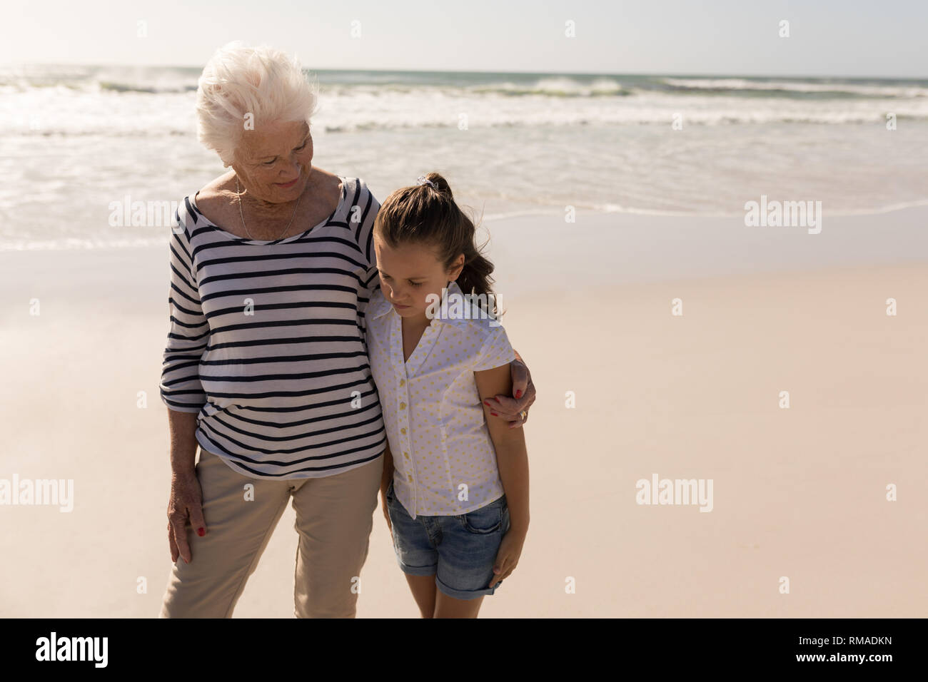 Senior woman and her granddaughter with arms around standing on beach Stock Photo