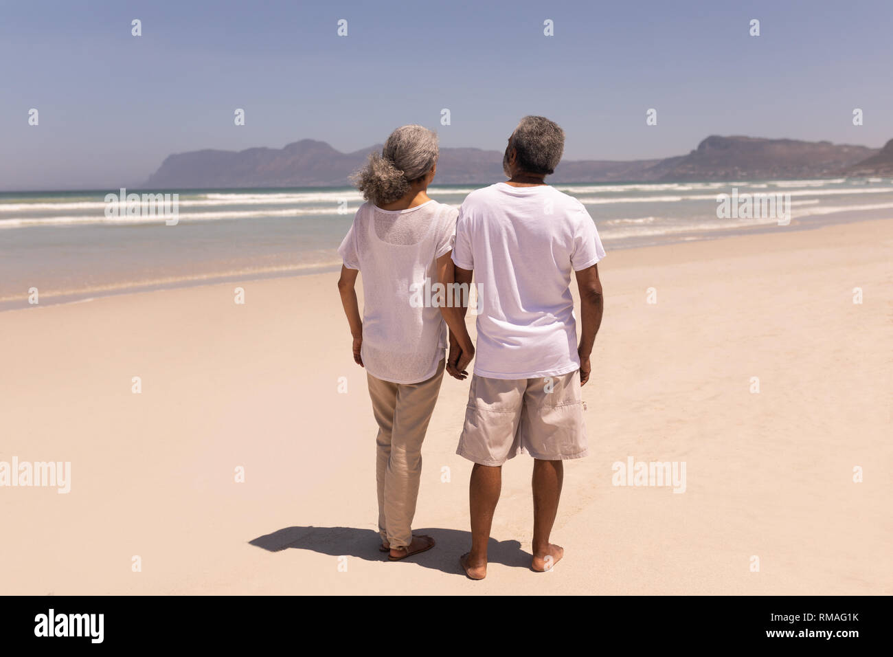 Senior couple holding hands and walking on beach with mountains in the background Stock Photo
