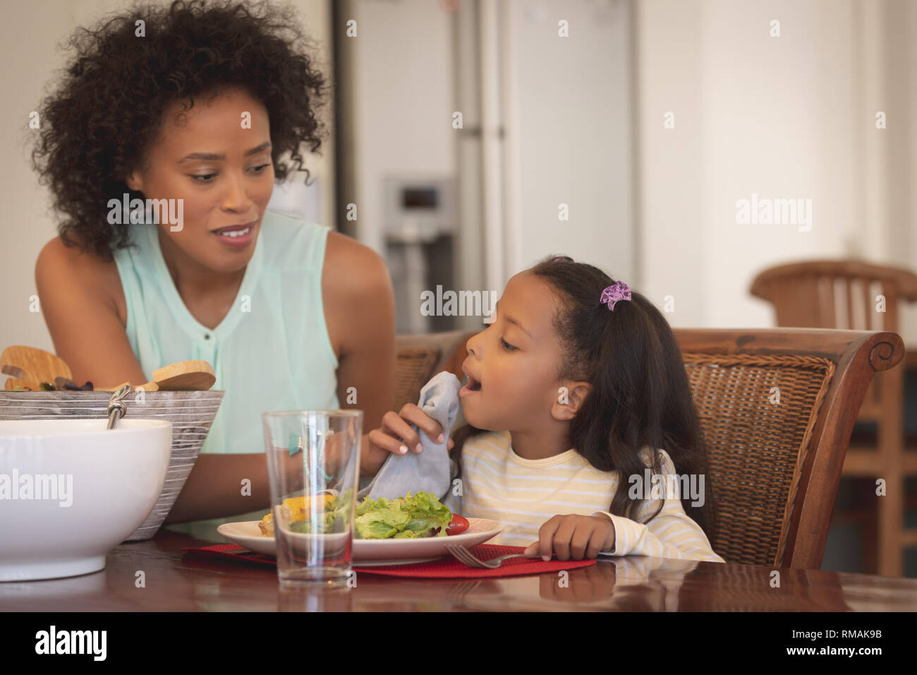 African American mother cleaning her daughter mouth with napkin at dining table Stock Photo