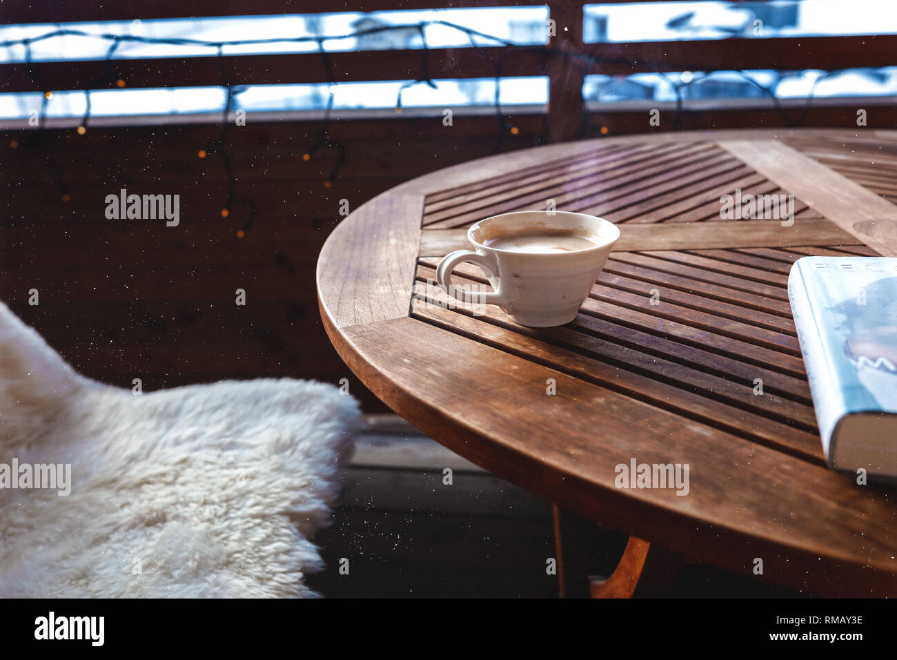 Wooden coffee table standing outdoor on the terrace, tasty aromatic cappuccino in a white cup, steam waving. Book lying near the cup. Bench covered Stock Photo