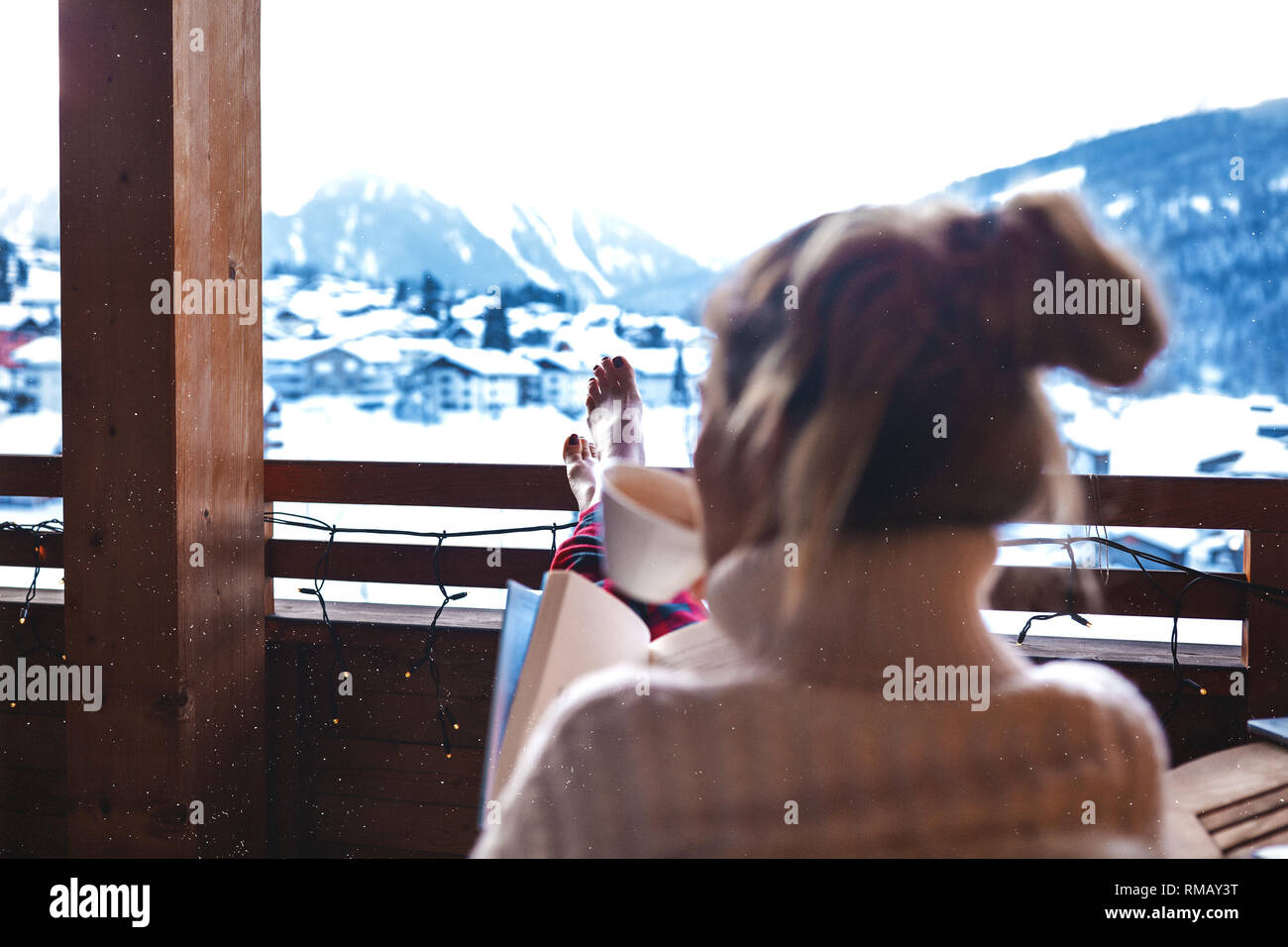 Pretty girl sitting on the wooden balcony in a hotel, put her long legs on the handrail and drinking morning latte. Wearing pajama and sweater. Enjoys Stock Photo