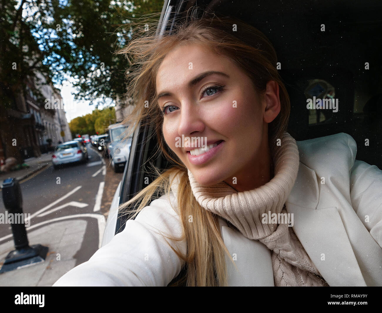 Portrait of beautiful young lady riding in a car, making selfie from the window on her phone. Smiling and cheerful girl in creamy sweater and white Stock Photo