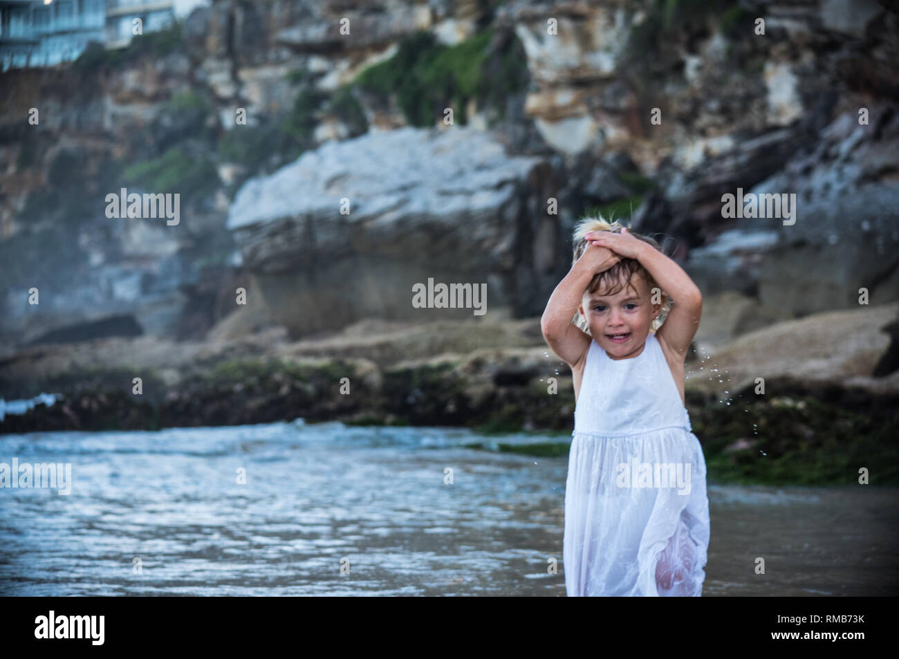 little blond baby girl wetting her hair and playing in the water of a beautiful empty beach with rocks Stock Photo